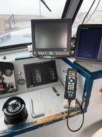 A close-up of a boat navigation console with dual screens, a handheld controller, a compass, and various control panels on a weathered blue and white dashboard.