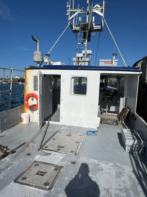 A white and blue fishing vessel cabin and deck area photographed outdoors under a clear blue sky, with metal hatch covers, rigging, and a life preserver visible.