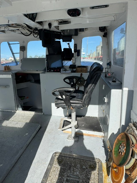 An interior view of a boat cabin with a black and gray captain's chair, computer monitors, navigation controls, and mechanical pulleys visible near the top right.