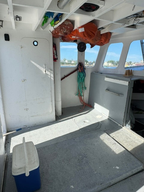 The interior cabin of a boat with white walls, windows showing blue sky and water, an orange life jacket hanging on the port side, coiled ropes, shelving, and a gray deck floor.