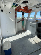 The interior cabin of a boat with white walls, windows showing blue sky and water, an orange life jacket hanging on the port side, coiled ropes, shelving, and a gray deck floor.