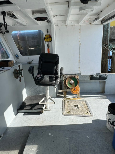 A black captain's chair with a headrest and armrests, positioned on a boat's deck next to a white cabin interior and gray hull.