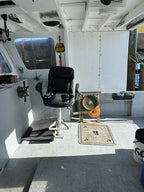 A black captain's chair with a headrest and armrests, positioned on a boat's deck next to a white cabin interior and gray hull.