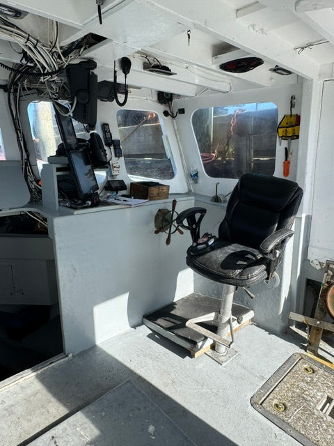 An overhead view of a boat's navigation bridge featuring a black captain's chair, steering wheel, radar screen, and various marine electronics mounted on a gray console table.