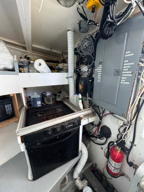 A close-up, high-angle view of a cluttered boat galley kitchen area, showing a black glass cooktop, electrical panel, vent pipes, and a fire extinguisher.