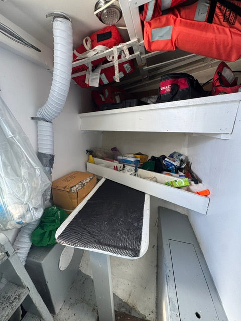A cluttered storage or utility space with white shelving and ventilation ducts. Visible items include orange and red life jackets or safety gear on the left, a black mesh panel leaning on a cardboard box, and various miscellaneous items in the center and right areas.