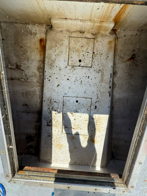 A dirty, stained metal utility compartment or storage box interior, showing rust, grime, and dirt accumulation on a light-colored floor with two square recessed areas.
