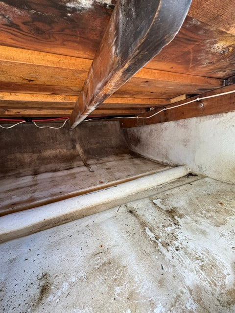 A close-up view of a rustic wooden wall with a large dark wooden beam or board in the foreground, a white pipe running vertically, and a concrete floor in a dim interior space.