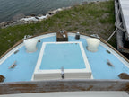 Top view of a boat's deck with a blue hatch and white cylindrical objects on a grassy area near water.