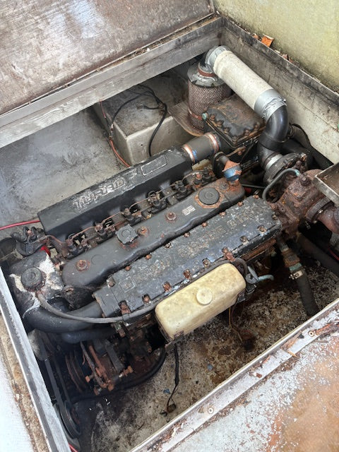 A used marine engine, identified as a Yanmar engine by the visible brand marking on the valve cover, shown inside its open engine compartment with rusted metal surfaces and various hoses and pipes.
