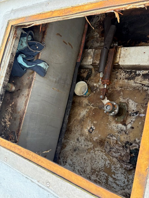 A close-up view of a heavily damaged, dirty mechanical compartment or opening with rusted metal trim, a hydraulic cylinder, concrete floor, and debris.