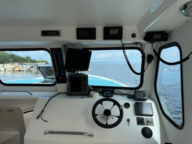The helm station of a boat with a white interior, featuring a black steering wheel, various electronic navigation screens, and a view of the open water and shoreline through the windows.