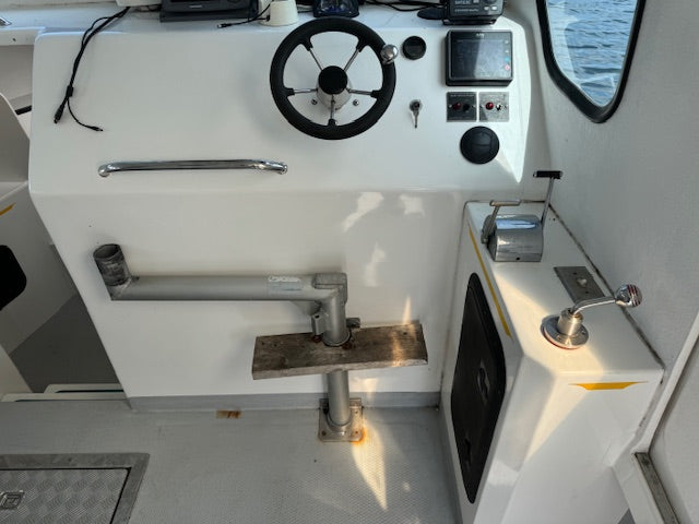 A close-up view of a boat's helm station featuring a white console wall, a black steering wheel, navigation screens, a metal grab handle, and a wooden deck attachment bracket.