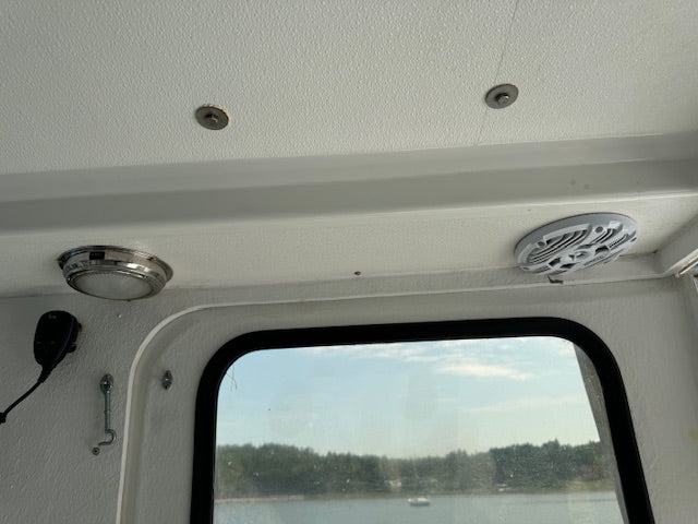 The interior ceiling of a boat cabin with a textured white ceiling, a circular dome light, a speaker, and a window looking out onto water and trees.