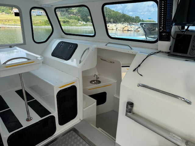 The interior of a boat cabin with white molded fiberglass surfaces, black storage compartments, chrome handles, and windows showing water, shoreline, and sky outside.