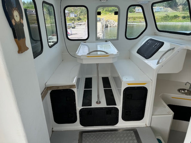 The cockpit interior of a white boat, featuring a central table, storage compartments with black panels, a helm station, and windows looking out onto water and green shoreline.