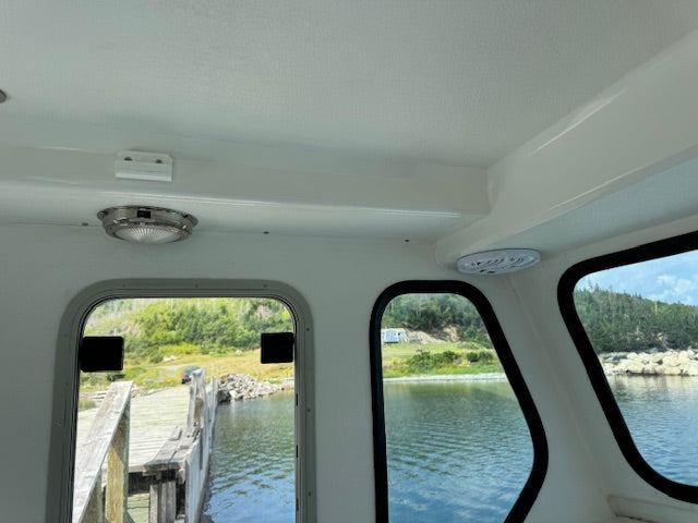 The interior cabin of a boat with white walls and ceiling, featuring arched windows looking out onto water and a forested shoreline.