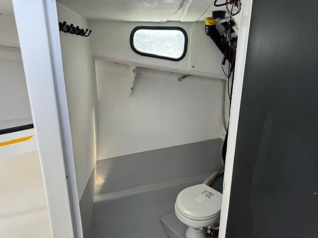 A compact white toilet with a closed lid installed in a small boat or RV bathroom with white walls, a gray floor, a small oval window, and visible wiring and storage hooks.