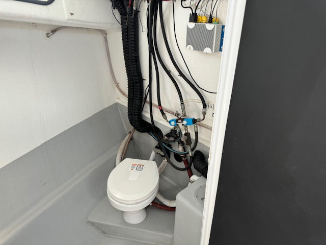 A compact white portable toilet with a black waste tank and connected plumbing pipes inside a white and gray enclosed space.