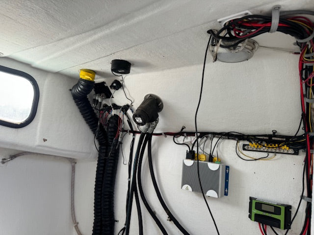 An overhead view of an electrical setup mounted on the white ceiling of a boat cabin, featuring various black cables, a black corrugated duct, a black camera or sensor, a gray electronic unit, and a green digital display.