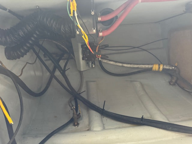 Close-up of an electrical or plumbing connection inside a boat or marine structure, showing black cables, a corrugated black hose, a red hose, and metal fittings on a light gray interior surface.