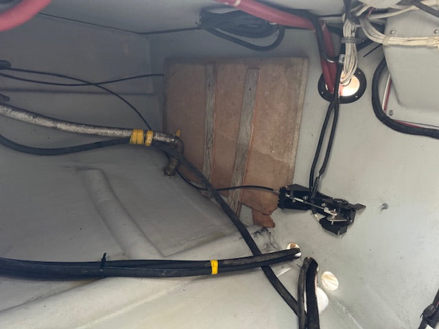 A close-up view of a boat's engine compartment showing a large brown foam battery with two vertical metal straps, surrounded by various black cables, wires, and hoses inside a light gray plastic compartment.