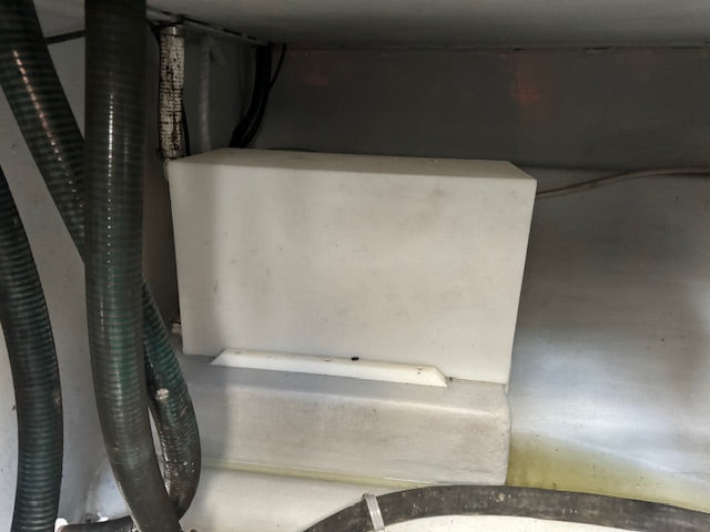 A white plastic utility box or filter component installed in a metallic silver machine compartment with dark corrugated hoses on the left.