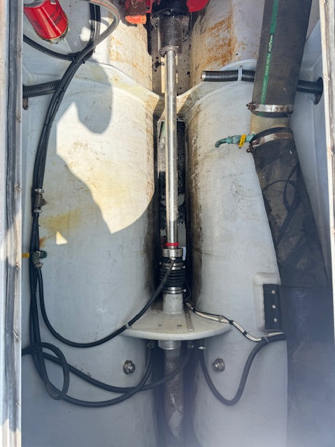 Close-up of a marine mechanical compartment showing a vertical metal pump or propeller shaft assembly between two large cylindrical tanks, with black hoses, a green pipe, and rusted metal surfaces.
