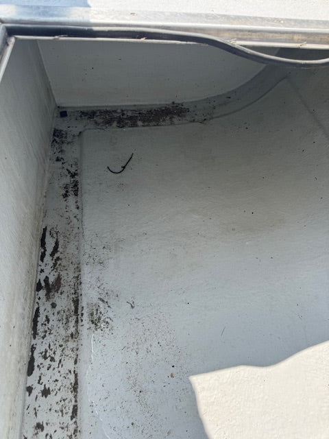 The inside corner of a white painted metal or plastic compartment, showing dirt, grime, and debris along the bottom edge and walls.
