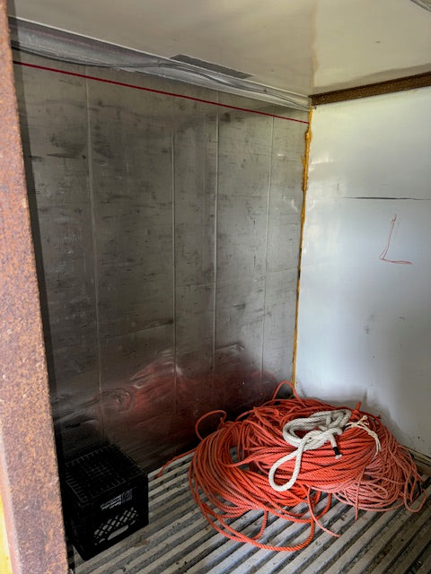A used industrial or utility room interior with a large transparent plastic sheet installed on a concrete wall, orange and white coiled cables on a metal floor, and a black plastic crate.