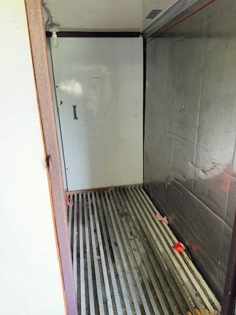 The interior of a commercial refrigerator or freezer compartment, showing a white door with a handle, a stainless steel slatted floor, and a textured metallic side wall.