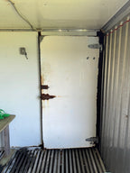 A white industrial freezer door with rust and wear, installed inside a metal shipping container or trailer.