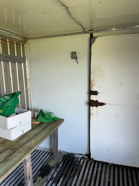 The interior of a rustic trailer or shed with a white door, metal slatted floor, and a wooden table holding white boxes and green bags.