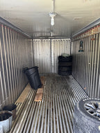 The interior of a cargo trailer showing a corrugated metal wall, slatted metal floor, a hanging light bulb, a black trash can, stacked tires, and a Road Grout tire in the foreground.