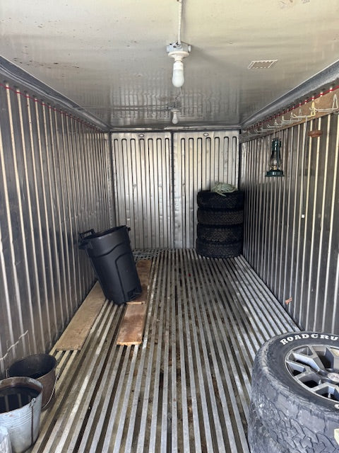The interior of a cargo trailer showing a corrugated metal wall, slatted metal floor, a hanging light bulb, a black trash can, stacked tires, and a Road Grout tire in the foreground.