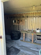 An unfinished workshop or garage interior with corrugated metal walls and floor, a wooden workbench, tools, a stack of tires, and an electrical panel mounted on the wall.