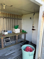 A cluttered storage or workshop space with corrugated metal walls and floor, a wooden workbench, white boxes, a green plastic bag, and a light blue trash bin in the foreground.