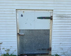 A weathered metal garage door with a white upper section and a textured gray lower section, set in a white-sided building with a concrete walkway.