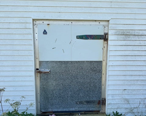 A weathered metal garage door with a white upper section and a textured gray lower section, set in a white-sided building with a concrete walkway.