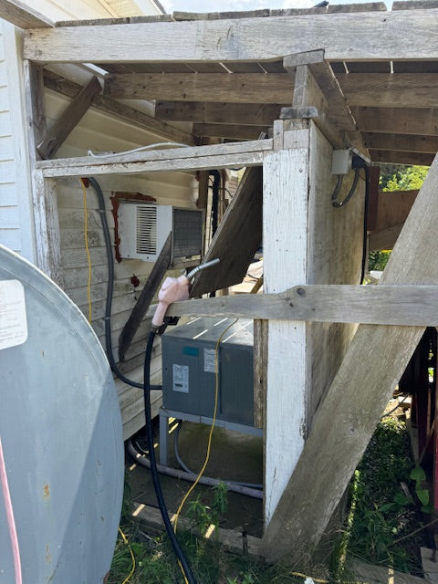 A residential outdoor air conditioner or heat pump unit housed under a weathered wooden porch structure, with a water heater unit visible to the left and a satellite dish edge in the foreground.