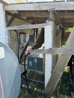 A residential outdoor air conditioner or heat pump unit housed under a weathered wooden porch structure, with a water heater unit visible to the left and a satellite dish edge in the foreground.