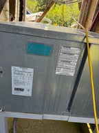A large galvanized steel HVAC air handler or condenser unit with visible KippLite branding and labels, photographed in a real workshop/outdoor setting with tools and greenery in the background.