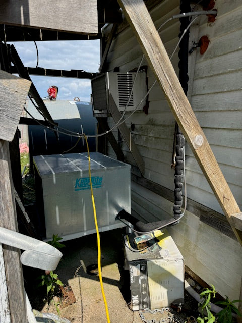 A large metal outdoor air conditioner or heat pump unit sits in a tight, cluttered outdoor space beside a house with white siding, wooden beams, exposed wiring, and a yellow cord running down the center.