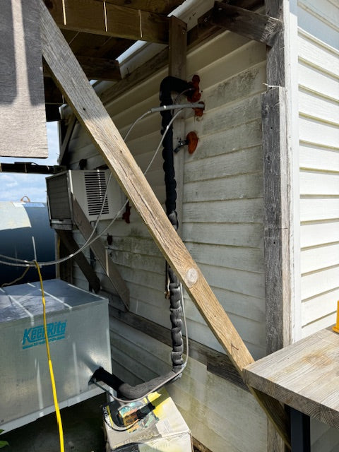 An outdoor HVAC or plumbing setup against a house wall, showing a black flexible duct, red valves, a silver metal box with blue text, and wooden support beams in a real environment.