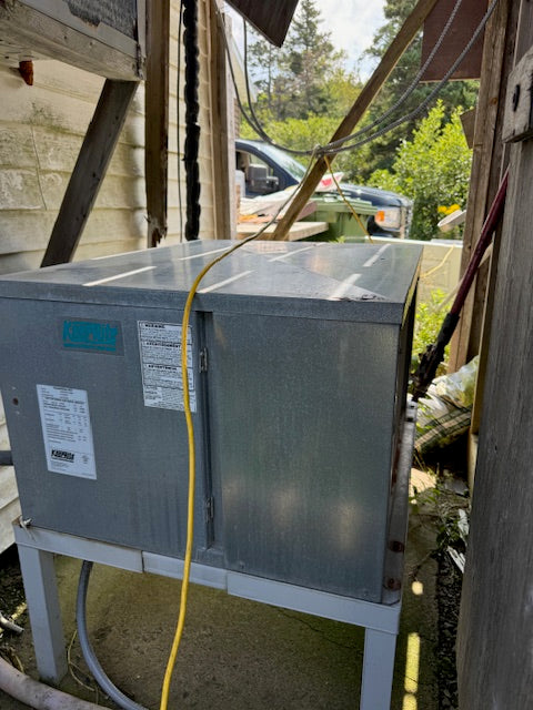 A large galvanized metal outdoor air conditioner or heat pump unit sitting on a metal support frame in a backyard or utility area, with a yellow power cord running down the front.