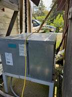 A large galvanized metal outdoor air conditioner or heat pump unit sitting on a metal support frame in a backyard or utility area, with a yellow power cord running down the front.
