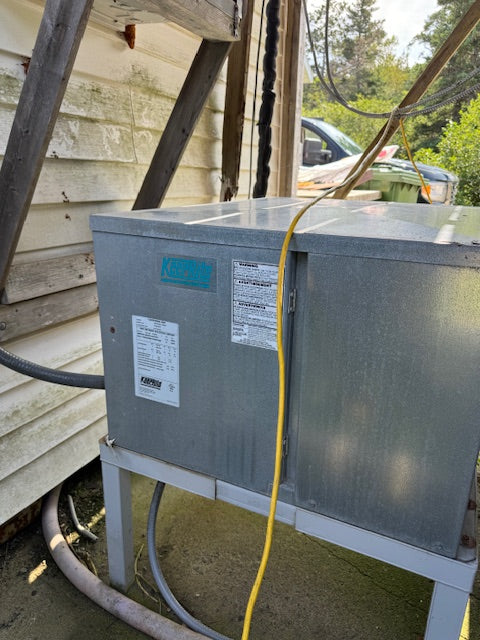 A large galvanized steel HVAC outdoor unit sitting on a metal support frame outdoors next to a house siding, with a yellow power cord running down the front.