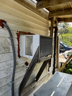 A white window air conditioner mounted on the exterior wall of a house, supported by wooden braces under a wooden porch overhang. The wall has peeling beige siding and rust-colored sealant around the unit.