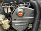 A close-up of a Yanmar marine engine with a large silver engine cover, black hoses, and filters visible in a tight mechanical bay.