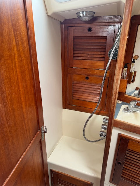 A close-up view of a compact bathroom vanity with dark brown wood louvered cabinets, a cream-colored bathtub/shower area, a flexible metal hose, and part of a sink and mirror visible on the right.